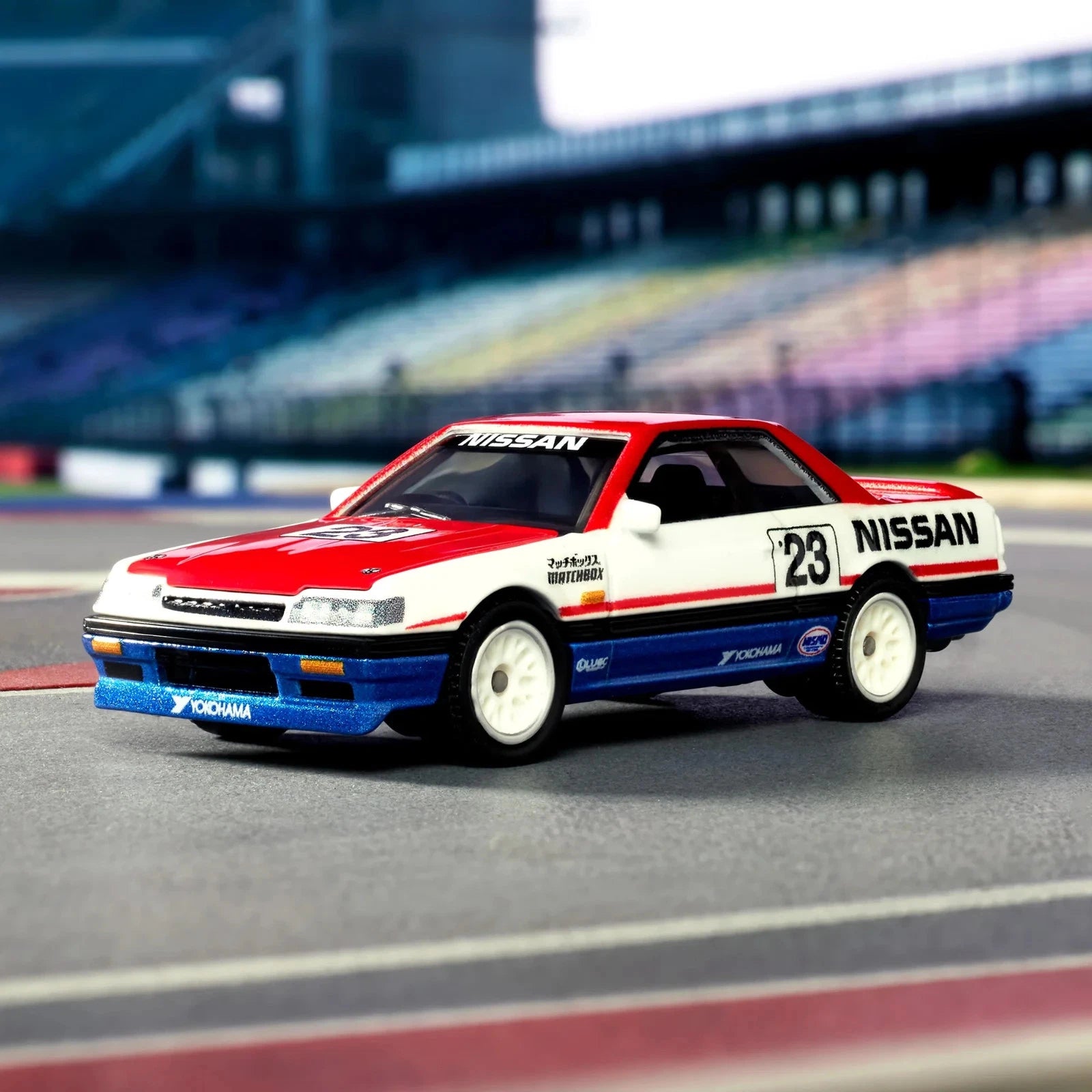 A die-cast model of a red, white, and blue Nissan race car with the number 23, positioned on a racetrack with blurred grandstands in the background.